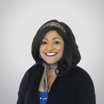 Annie Varughese MD vilkintis juodą smokingą ir varlytę, stovi priešais banerį su logotipais, įskaitant Oscars ir HeartDaHeart Foundation, oficiali renginio aplinka, neutralus ir profesionalus įspūdis, matomas tekstas: Oscars HeartDaHeart Foundation UnmaskHeartDisease