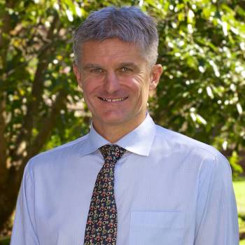 Gordon Crozier DO vilkintis juodą smokingą ir varlytę, stovi priešais banerį su logotipais, įskaitant Oscars ir HeartDaHeart Foundation, oficiali renginio aplinka, neutralus ir profesionalus įspūdis, matomas tekstas: Oscars HeartDaHeart Foundation UnmaskHeartDisease