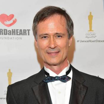 Douglas S Harrington MD vilkintis juodą smokingą ir varlytę, stovi priešais banerį su logotipais, įskaitant Oscars ir HeartDaHeart Foundation, oficiali renginio aplinka, neutralus ir profesionalus įspūdis, matomas tekstas: Oscars HeartDaHeart Foundation UnmaskHeartDisease