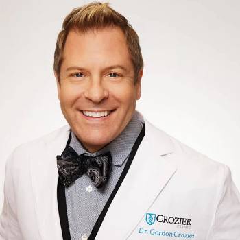 Gordon Crozier DO vilkintis juodą smokingą ir varlytę, stovi priešais banerį su logotipais, įskaitant Oscars ir HeartDaHeart Foundation, oficiali renginio aplinka, neutralus ir profesionalus įspūdis, matomas tekstas: Oscars HeartDaHeart Foundation UnmaskHeartDisease