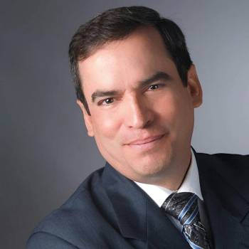 Luis Miguel Valdes MD vilkintis juodą smokingą ir varlytę, stovi priešais banerį su logotipais, įskaitant Oscars ir HeartDaHeart Foundation, oficiali renginio aplinka, neutralus ir profesionalus įspūdis, matomas tekstas: Oscars HeartDaHeart Foundation UnmaskHeartDisease