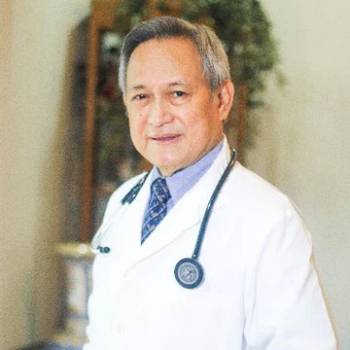 Gordon Crozier DO vilkintis juodą smokingą ir varlytę, stovi priešais banerį su logotipais, įskaitant Oscars ir HeartDaHeart Foundation, oficiali renginio aplinka, neutralus ir profesionalus įspūdis, matomas tekstas: Oscars HeartDaHeart Foundation UnmaskHeartDisease